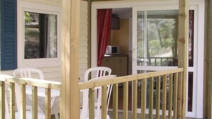 A wooden deck with white chairs, a small table, and a sliding glass door leading to an interior space with a red curtain and a microwave.