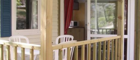 A wooden deck with white chairs, a small table, and a sliding glass door leading to an interior space with a red curtain and a microwave.