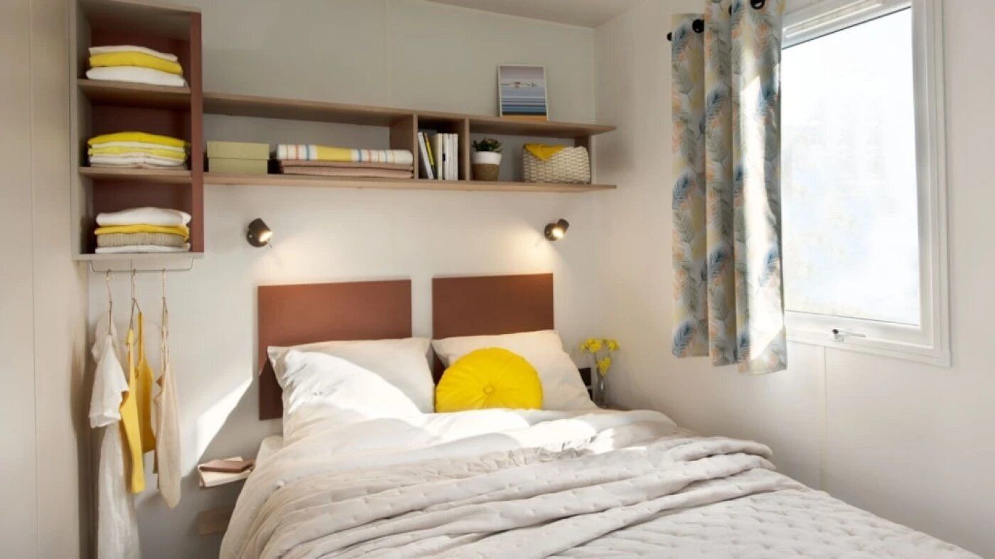 Een keurig opgeruimde slaapkamer met een bed, planken met opgevouwen handdoeken en een raam met gordijnen.