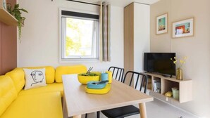 Un sofá amarillo brillante con un cojín, una mesa de madera con una ensalada en un cuenco, un televisor sobre un mueble bajo y una ventana con cortinas.