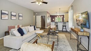 A modern living room with a white sofa, a coffee table, and a ceiling fan. There is a kitchen area visible in the background with a dining table and chairs. - Basketball Court, Pickleball, Karaoke, 6 Beds (Minneapolis)