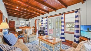 A spacious living room with wooden beams, a dining area, and a television displaying a tropical beach scene.