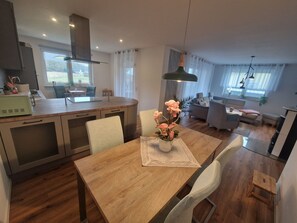 Eine moderne Küche mit Kochinsel, Essbereich mit Holztisch und Stühlen sowie ein Wohnzimmer mit Sofa und Couchtisch.