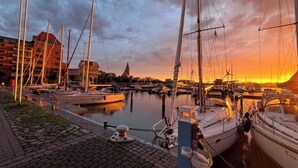 A marina with several boats docked, a cobblestone walkway, and buildings in the background.
