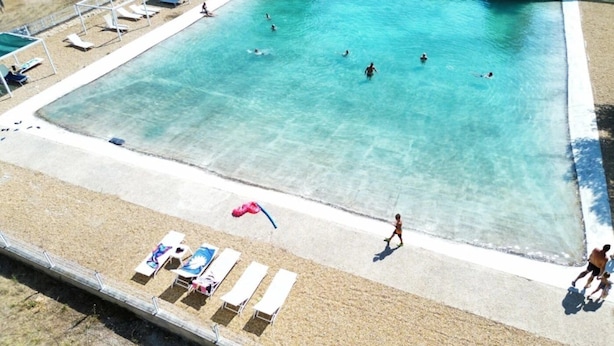 Una gran piscina con agua cristalina azul, rodeada por una playa de arena con sillas de descanso y sombrillas.