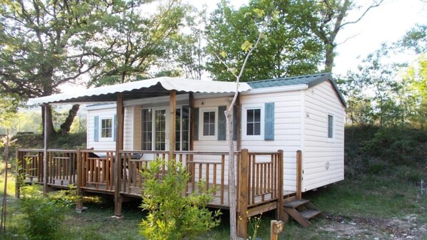 A small, single-story cabin with a porch and wooden railings, surrounded by trees.