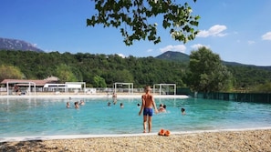 A swimming pool with people in the water, surrounded by a pebble beach and trees. - Camping 3 étoiles - Piscine  - ccbaehe (Mézel)