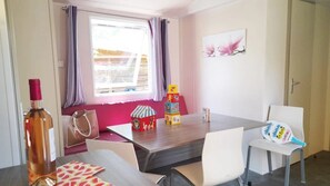 Quarto com mesa de madeira, cadeiras brancas, uma garrafa de vinho e janela com cortinas.