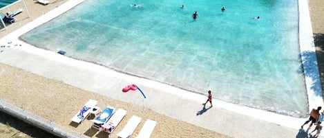 Una gran piscina con agua cristalina azul, rodeada por una playa de arena con sillas de descanso y sombrillas.