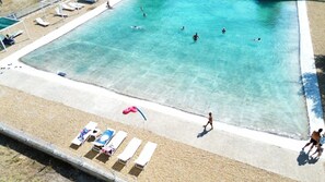 Uma grande piscina com água azul cristalina, rodeada por uma praia arenosa com espreguiçadeiras e guarda-sóis.