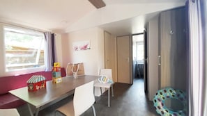 A room with a dining table, chairs, a window with curtains, a door, and a small play area with a colorful toy. - Camping 3 étoiles - Piscine  - ccbaehe (Mézel)
