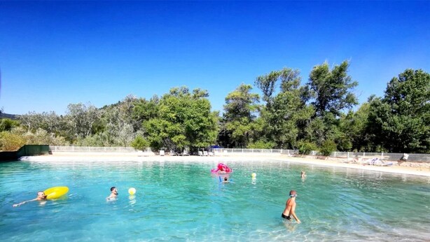 A swimming pool with clear blue water, surrounded by trees and a sandy beach area.