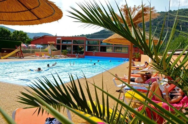 Una piscina con gente nadando y tomando el sol, rodeada de palmeras y un edificio de techo rojo.