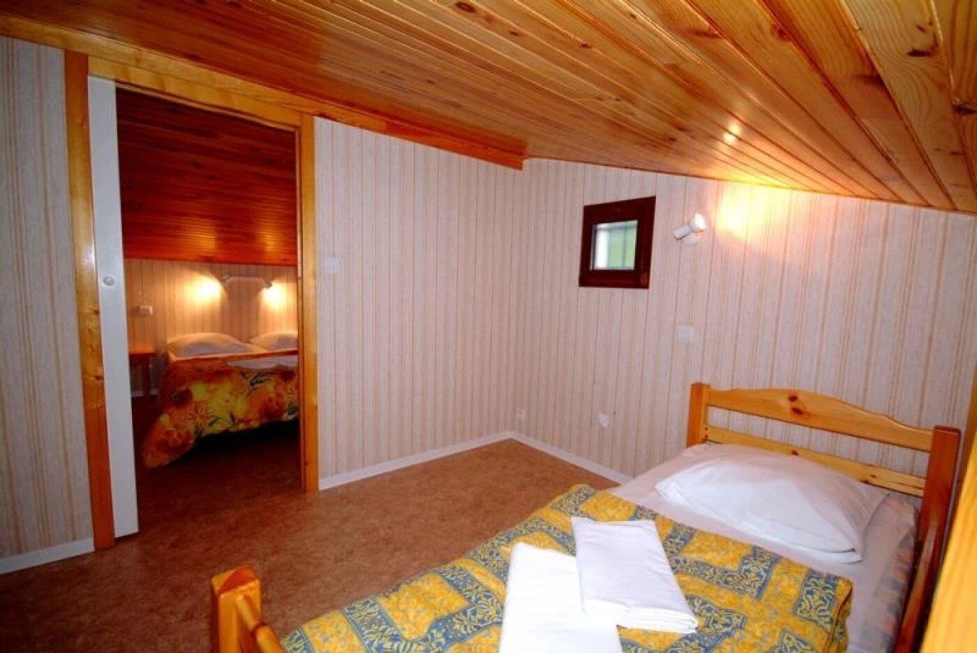 A bedroom with a wooden ceiling, a bed with a patterned bedspread, a small window, and a door leading to another room.