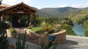 A wooden reception building with a glass door, surrounded by a stone wall and a wooden deck, overlooking a river and mountains. - ZEcamping 4* familial piscine locatif TV clim 27m2 4 pers (Saint-Rome-de-Tarn)