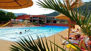A swimming pool area with lounge chairs and umbrellas, surrounded by palm trees and a building with large windows. - ZEcamping 4* piscine animations locatif TV clim 32m2 4 pers (Saint-Rome-de-Tarn)