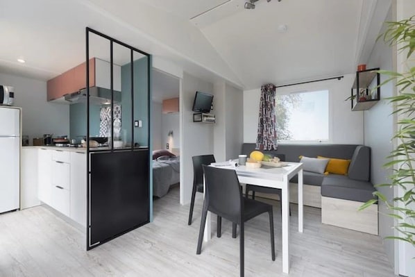 A modern kitchen with a black-framed sliding door, a dining area with a white table and black chairs, and a grey sofa with yellow cushions.