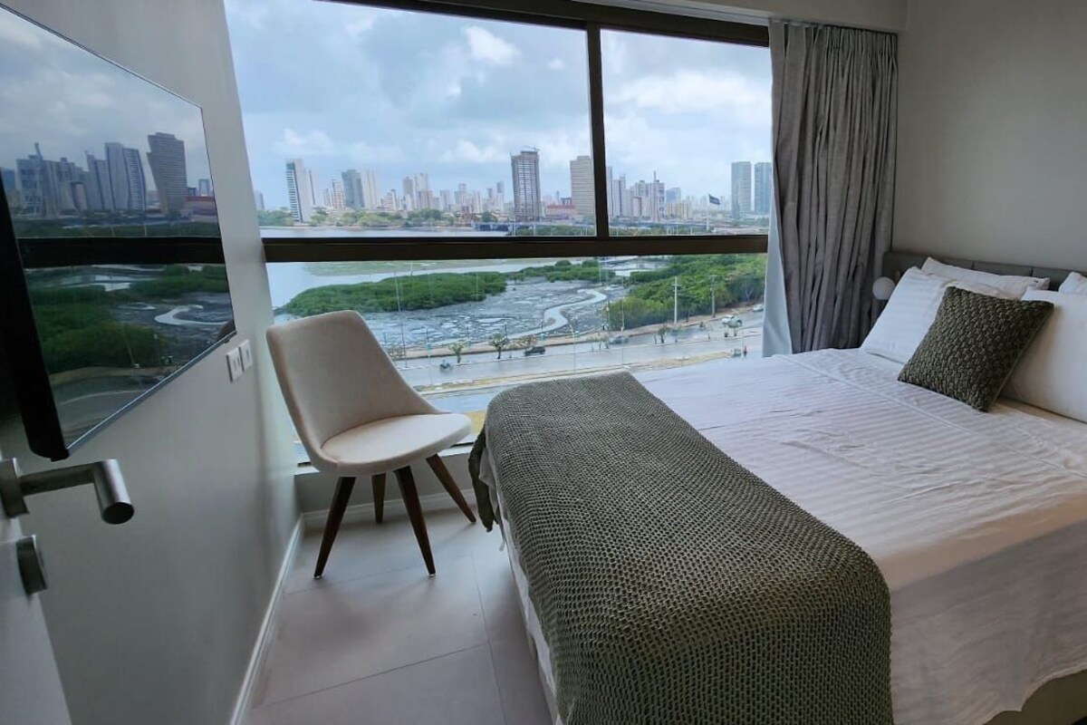 A bedroom with a large bed, a chair, and a view of a cityscape through a window.