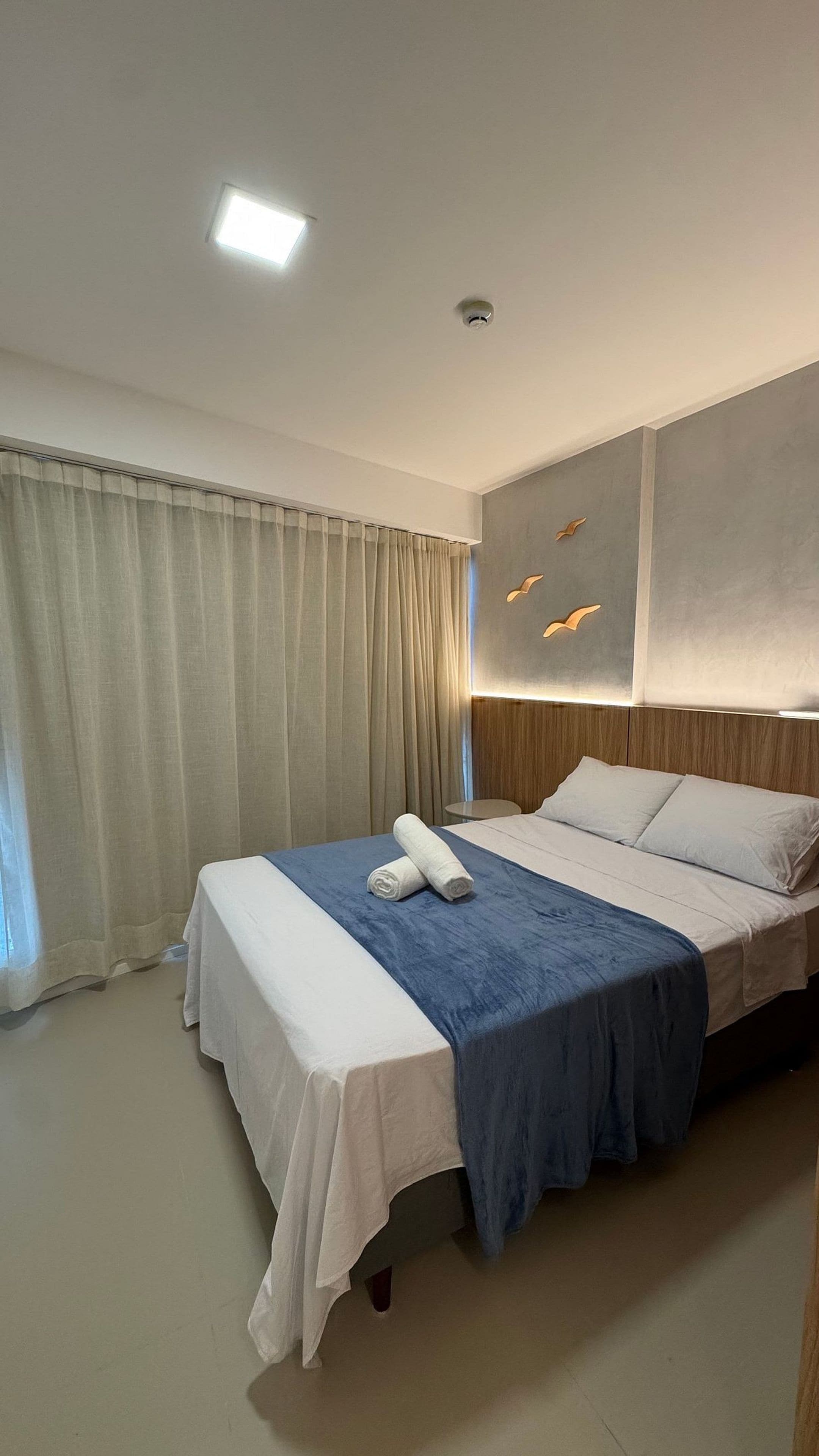 Una habitación de hotel con cama, cortinas, cabecera con luces y una lámpara de techo.