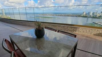 Una terrazza con tavolo e sedie, una pianta in vaso e un parapetto in vetro che si affaccia su un panorama urbano e una distesa d'acqua.