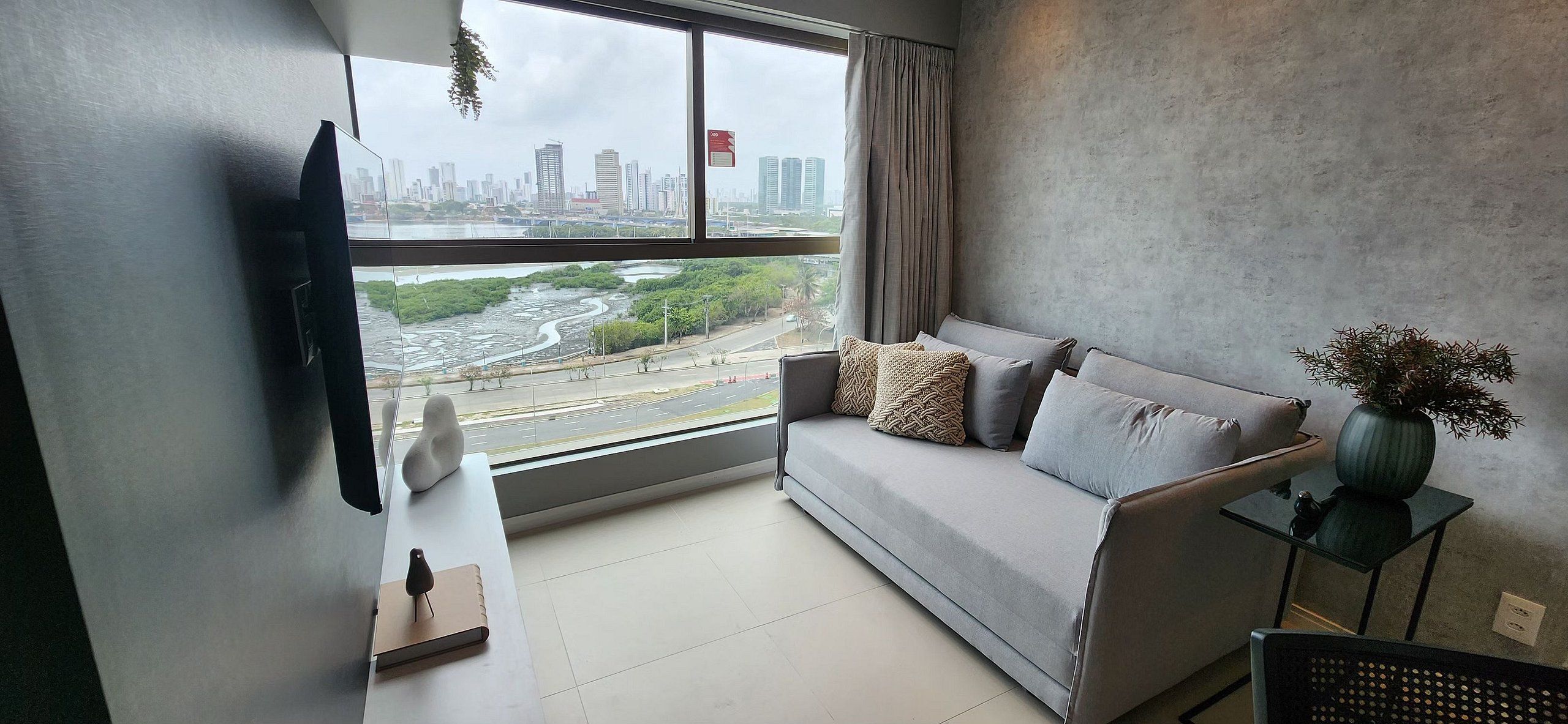 A modern living room with a grey sofa, a flat-screen TV mounted on the wall, and a large window offering a view of a cityscape and a river.