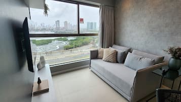A modern living room with a grey sofa, a flat-screen TV mounted on the wall, and a large window offering a view of a cityscape and a river.