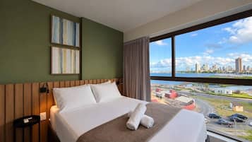 A hotel room with a bed, a nightstand, a lamp, and a window with a city view.