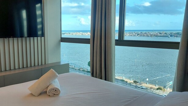 A hotel room with a bed, a rolled white towel, a TV on the wall, and a view of the sea and cityscape through large windows. - Flats no Parque do Cais Recife  (Recife)