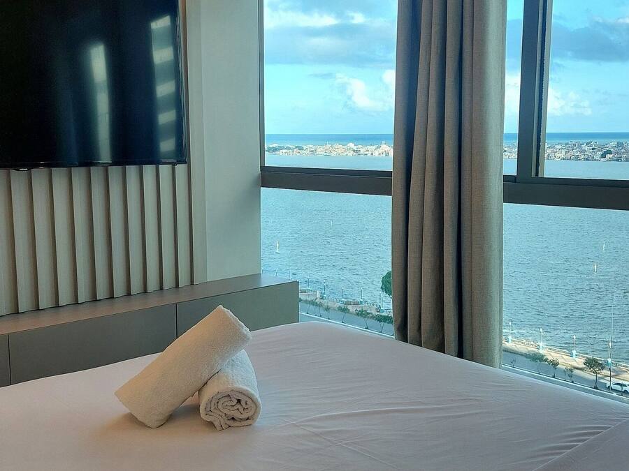 Quarto de hotel com uma cama, uma toalha branca enrolada, uma televisão na parede e vista para o mar e para a cidade através de grandes janelas.