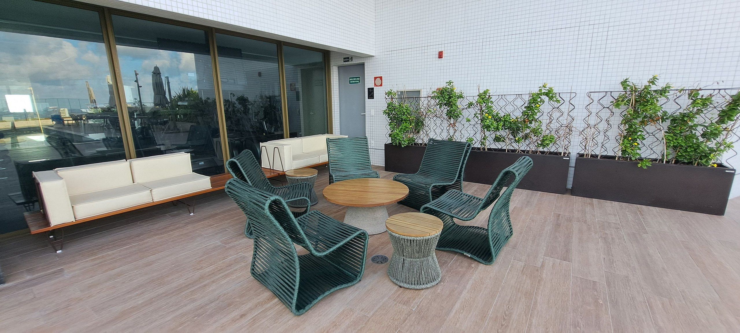 Un espacio exterior moderno con sillas de mimbre, una mesa redonda, un sofá blanco y una pared de vidrio con vista a la ciudad.