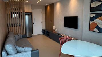 A modern living room with a sectional sofa, a dining table with chairs, a flat-screen TV mounted on the wall, and a kitchen area with a refrigerator.