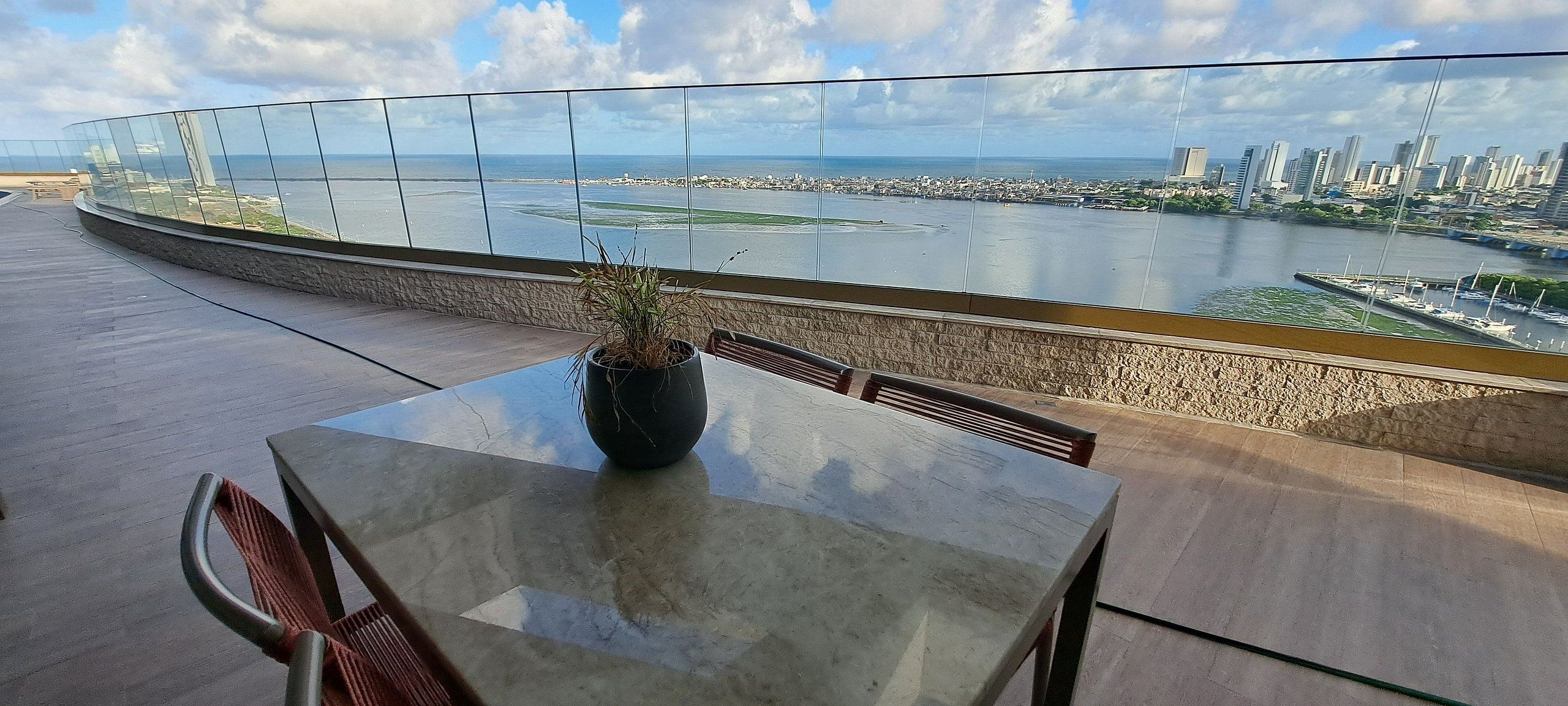 Terras met tafel en stoelen, een potplant en een glazen hekwerk met uitzicht op een stadsbeeld en een waterpartij.
