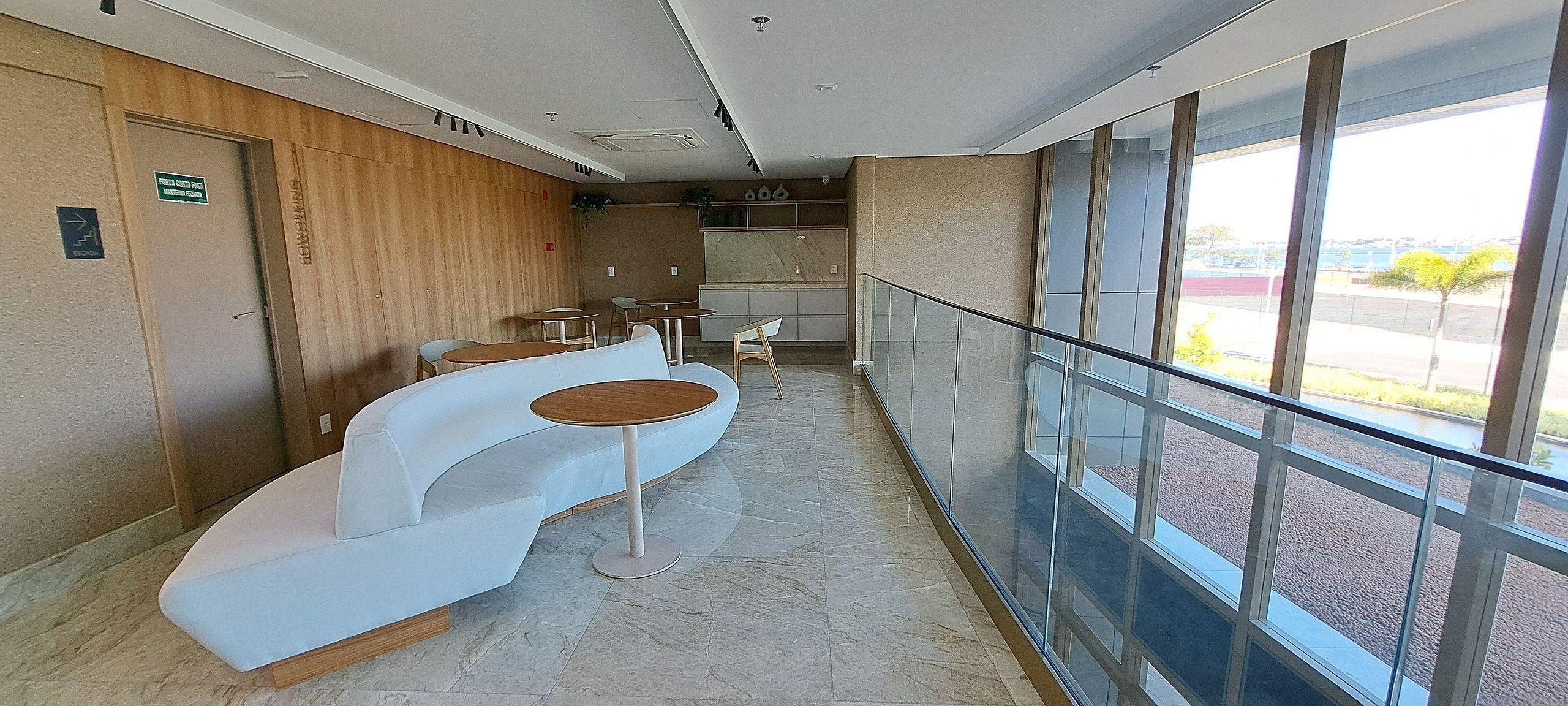 Eine moderne Hotelhalle mit einer gebogenen weißen Couch, kleinen runden Tischen und einem Glasgeländer mit Blick auf einen Parkplatz.