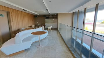 Un moderno vestíbulo de hotel con un sofá blanco curvo, pequeñas mesas redondas y una barandilla de vidrio que da a un aparcamiento.