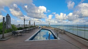 A rooftop pool area with lounge chairs and tables, overlooking a body of water and cityscape. - Flats no Parque do Cais Recife  (Recife)