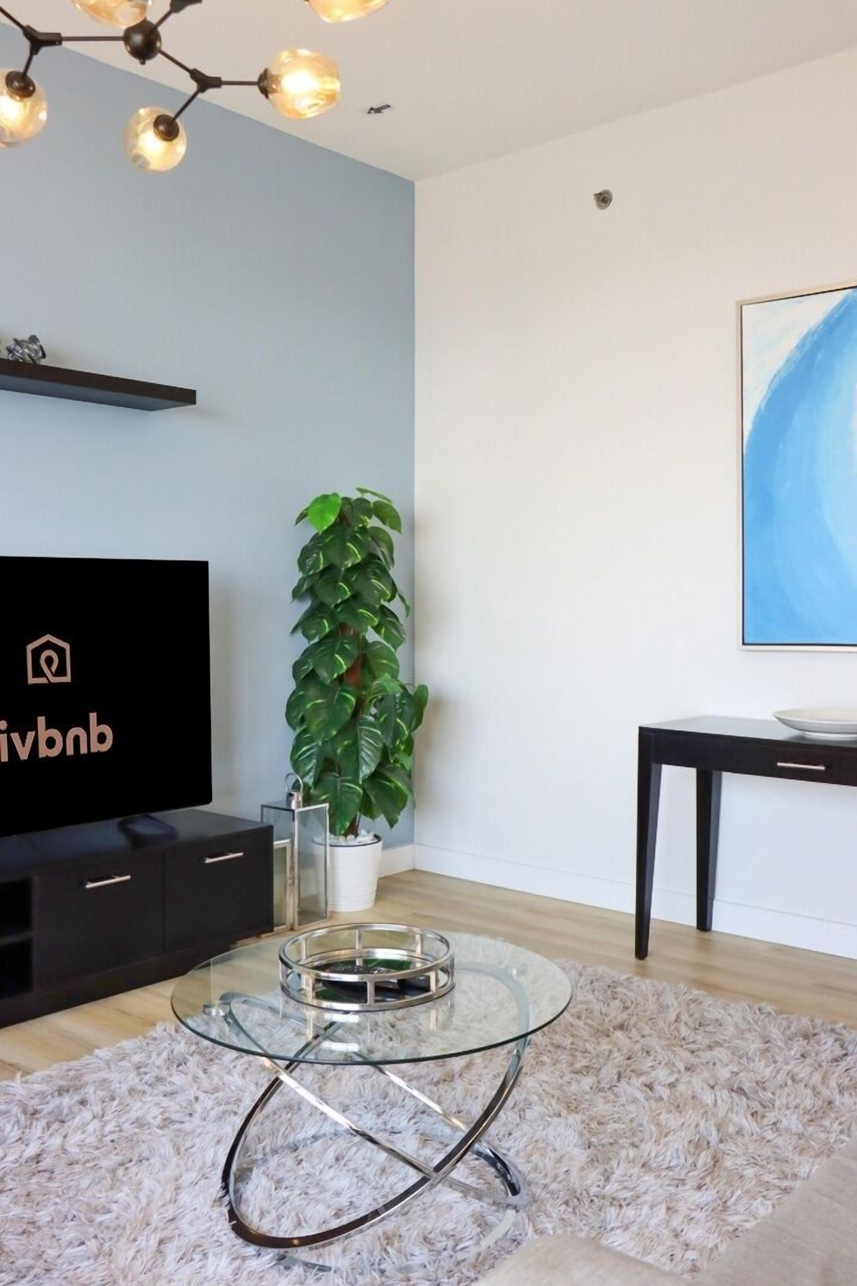 A modern living room with a flat-screen TV, a glass coffee table, a potted plant, and a painting on the wall.