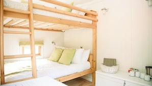 Ein Etagenbett mit Holzrahmen, einer weißen Matratze und grünen Kissen. Auf dem Nachttisch steht ein weißer Korbwannenkorb, ein weißer Schrank mit einem Wasserkocher und ein Fenster mit Vorhang.