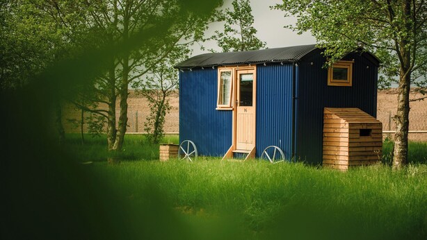Ein kleines, eingeschossiges Holzhaus mit Tür und Fenster, umgeben von Gras und Bäumen.