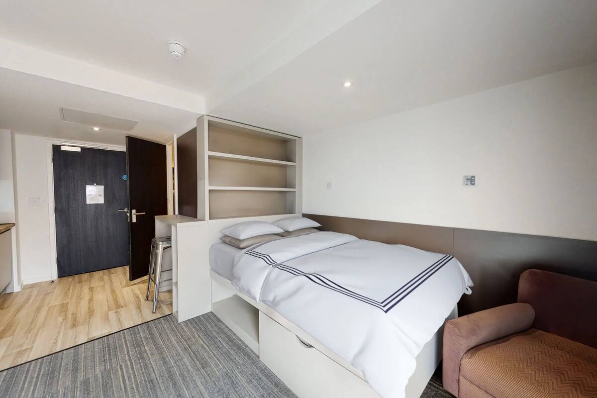 A compact hotel room with a single bed, a small seating area, and a door leading to a hallway.