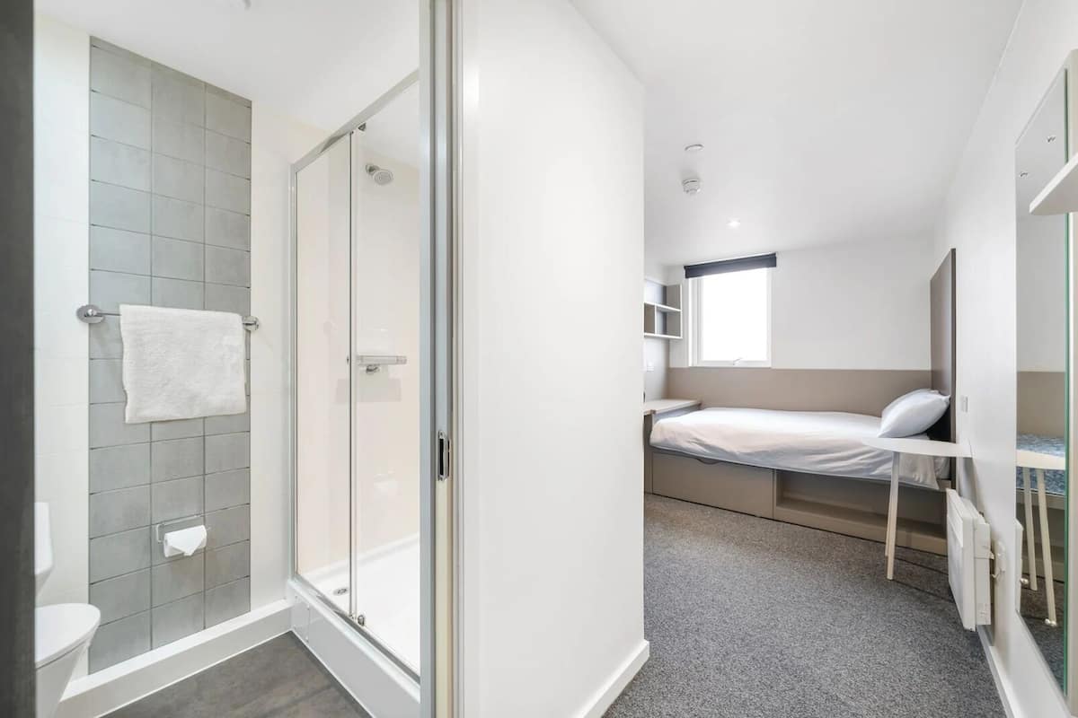 A compact bedroom with a shower, a bed, and a small window.