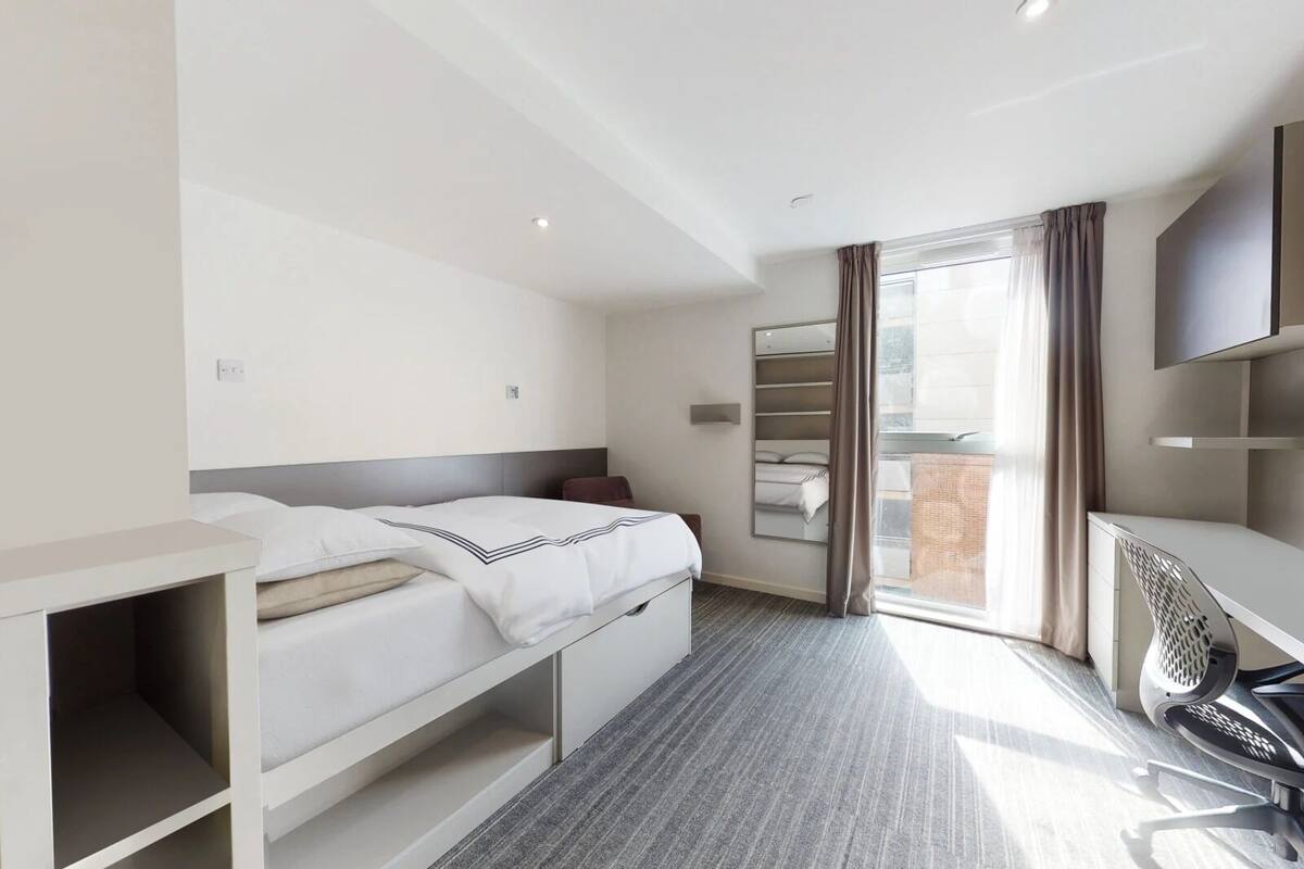 A hotel room with a bed, a desk, a chair, a TV, a window with curtains, and a balcony.