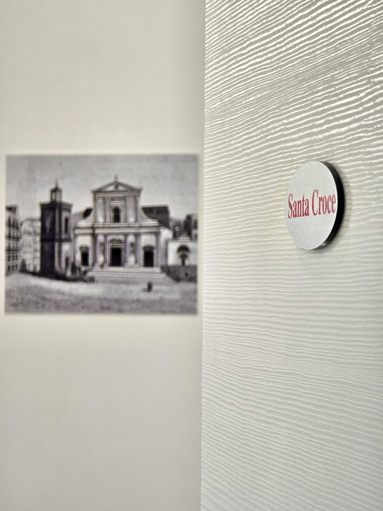 A black and white photograph of a historical building with a prominent central dome and a smaller tower to the left. A round button with the text "Santa Croce" is visible on the right side of the image.