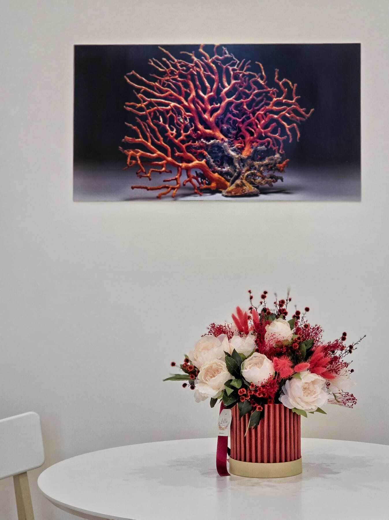 A round table with a vase of flowers and a painting of coral on the wall behind.