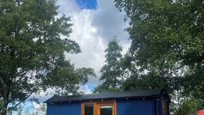 Une petite cabane bleue avec une porte et une fenêtre en bois, une table de pique-nique et deux grandes roues à l’extérieur.