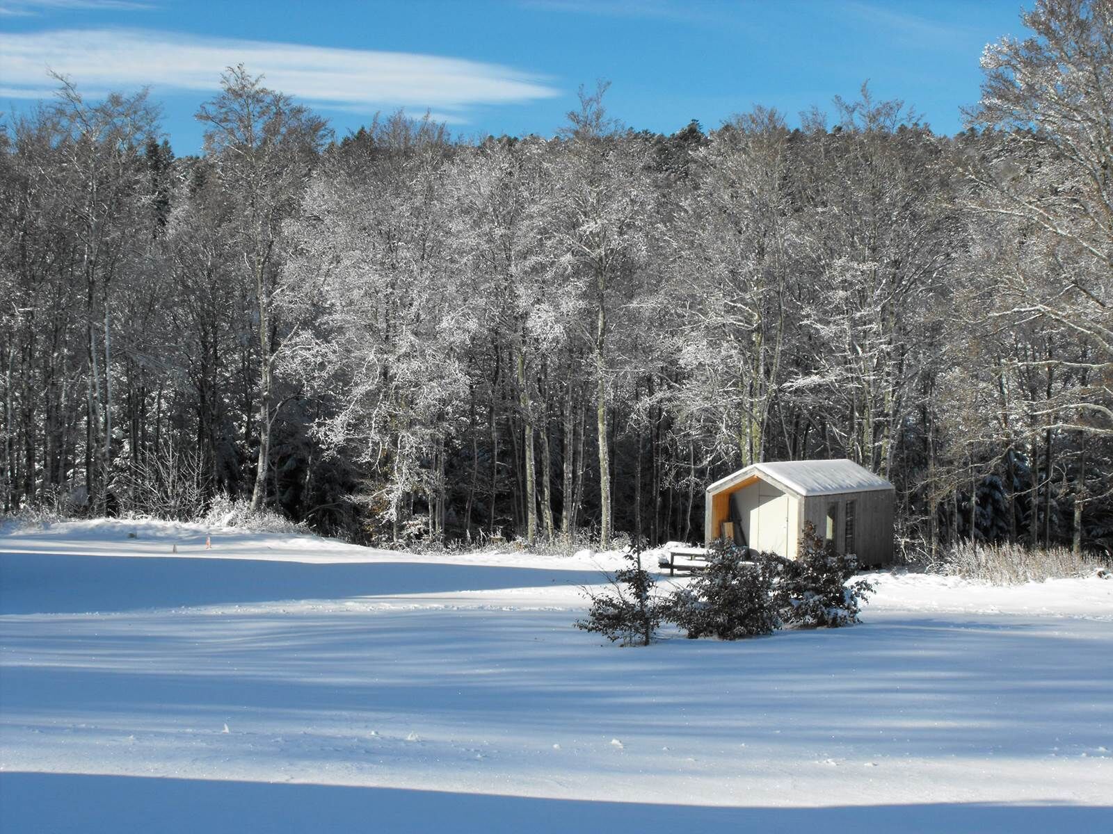 Une petite cabane en bois, d’un seul étage, avec un toit en pente, entourée d’un paysage enneigé et d’une forêt dense.