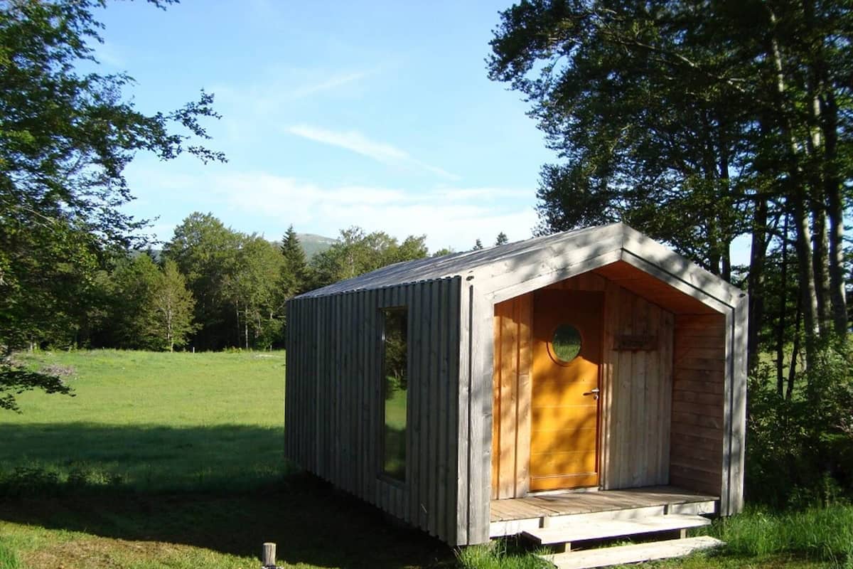 A small wooden cabin with a single door and a small porch, surrounded by a grassy field and trees.