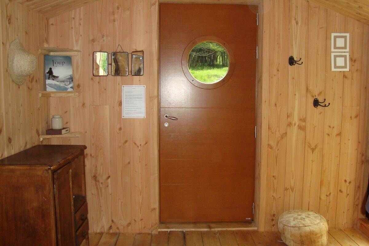 A wooden interior with a round door, a small stool, a shelf with a framed picture, and a wooden cabinet.