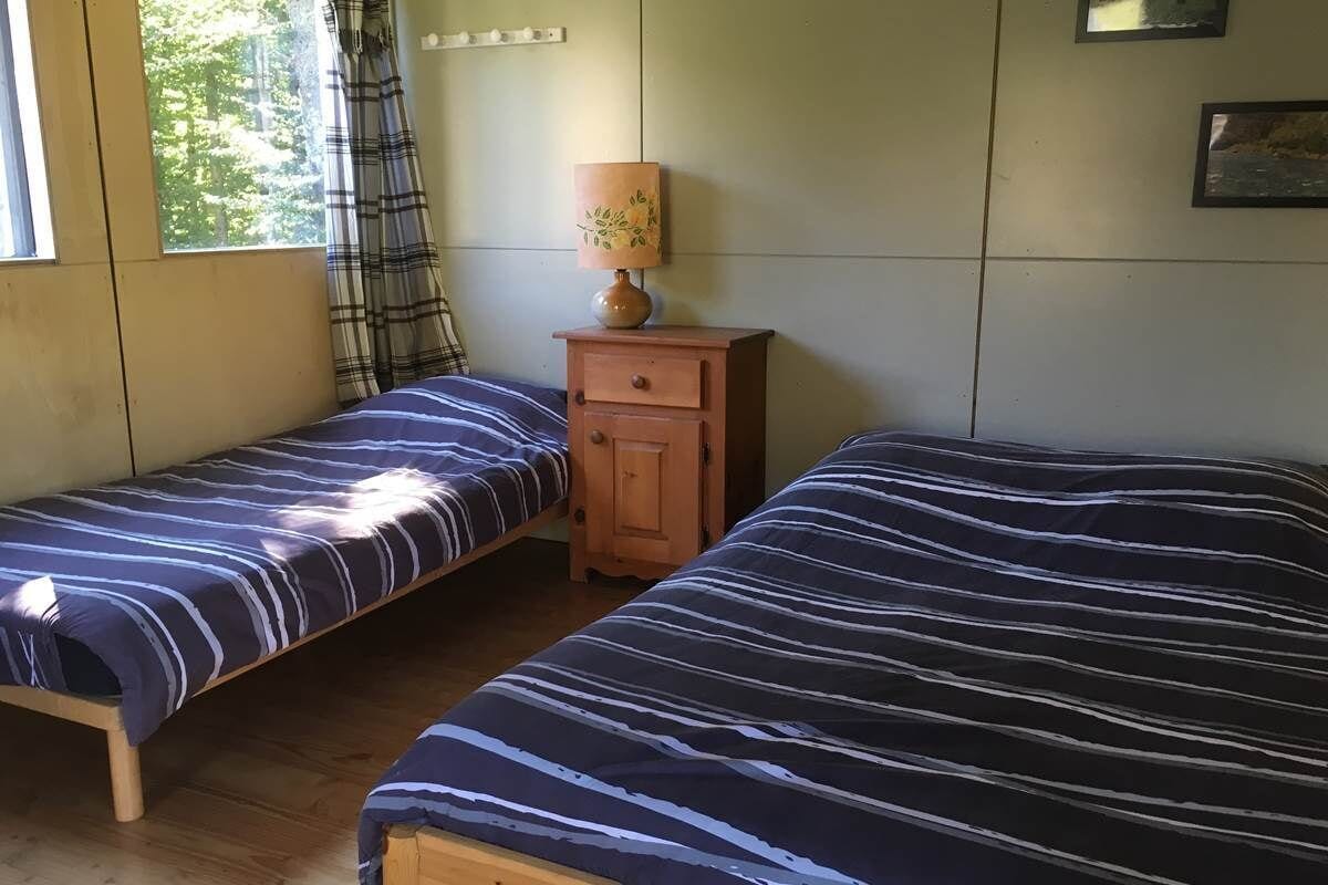 Et værelse med to senge, et lille trænatbord med en lampe, et ternet gardin og et vindue med udsigt til træer.