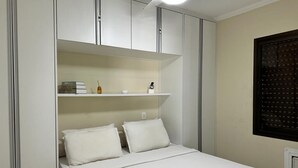 A bedroom with a bed, white walls, a ceiling fan, and built-in shelving with books and a small vase. - Flat Golden Beach (Centro Guarujá)