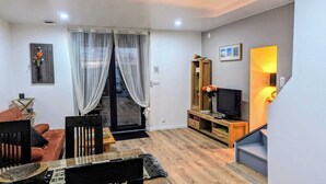 A living room with a sofa, a TV on a wooden stand, a dining table with chairs, and a staircase with a lamp. - Domaine la Griffe - Maison Mitoyenne Cou (La Roche-sur-Foron)